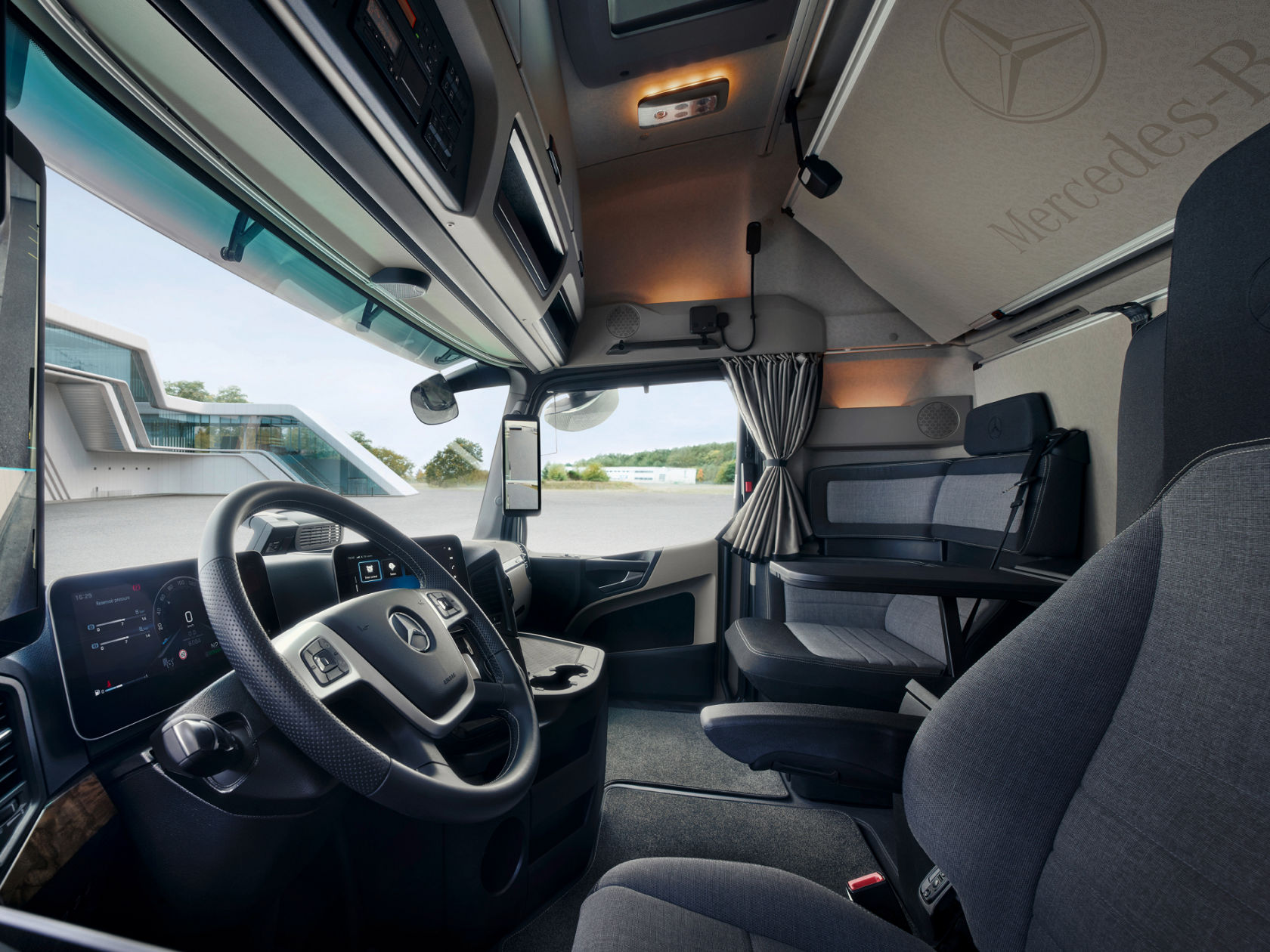 The image shows the spacious and comfortable interior of a modern Mercedes-Benz truck. You can see the steering wheel, the digital displays and the living area with a table and seating, which emphasises the functionality and comfort for long-distance journeys.