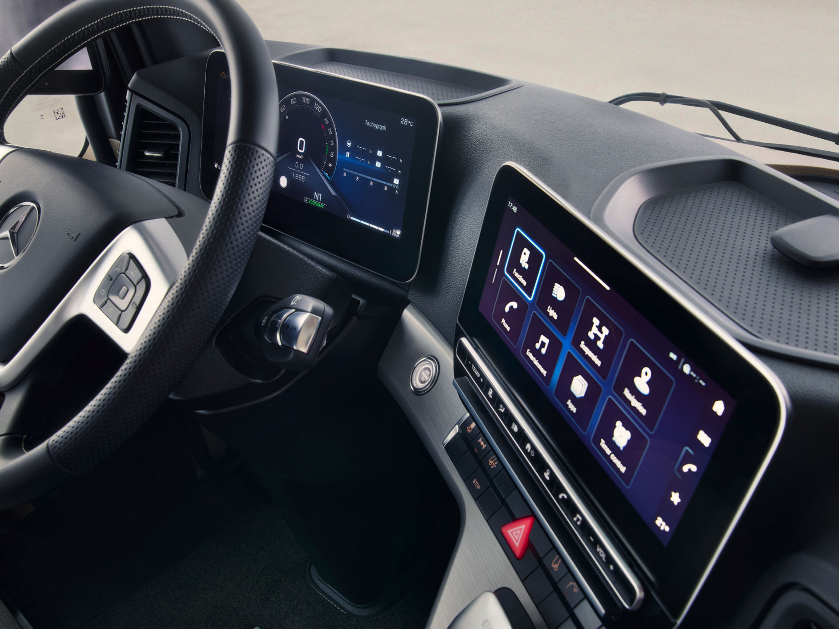 The image shows the modern interior of a Mercedes-Benz truck, with the focus on the steering wheel and the digital displays. There is a digital instrument cluster behind the steering wheel and a large touchscreen in the centre console, which is displaying various functions such as navigation and entertainment.