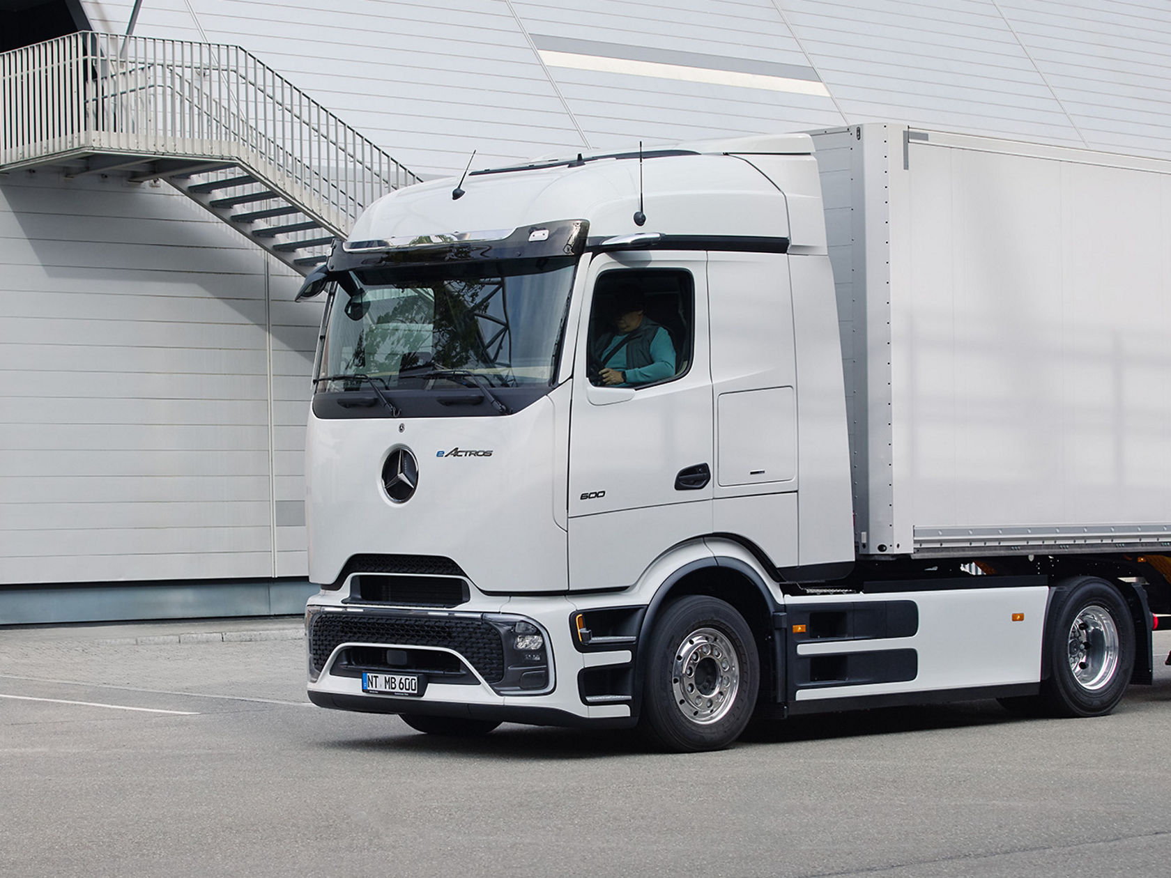 A white eActros with ProCabin from Mercedes-Benz Trucks with a box body drives on tarmac, while in the background you can see a modern building with an outside staircase and a large ventilation pipe. A driver is visible in the cab of the truck.