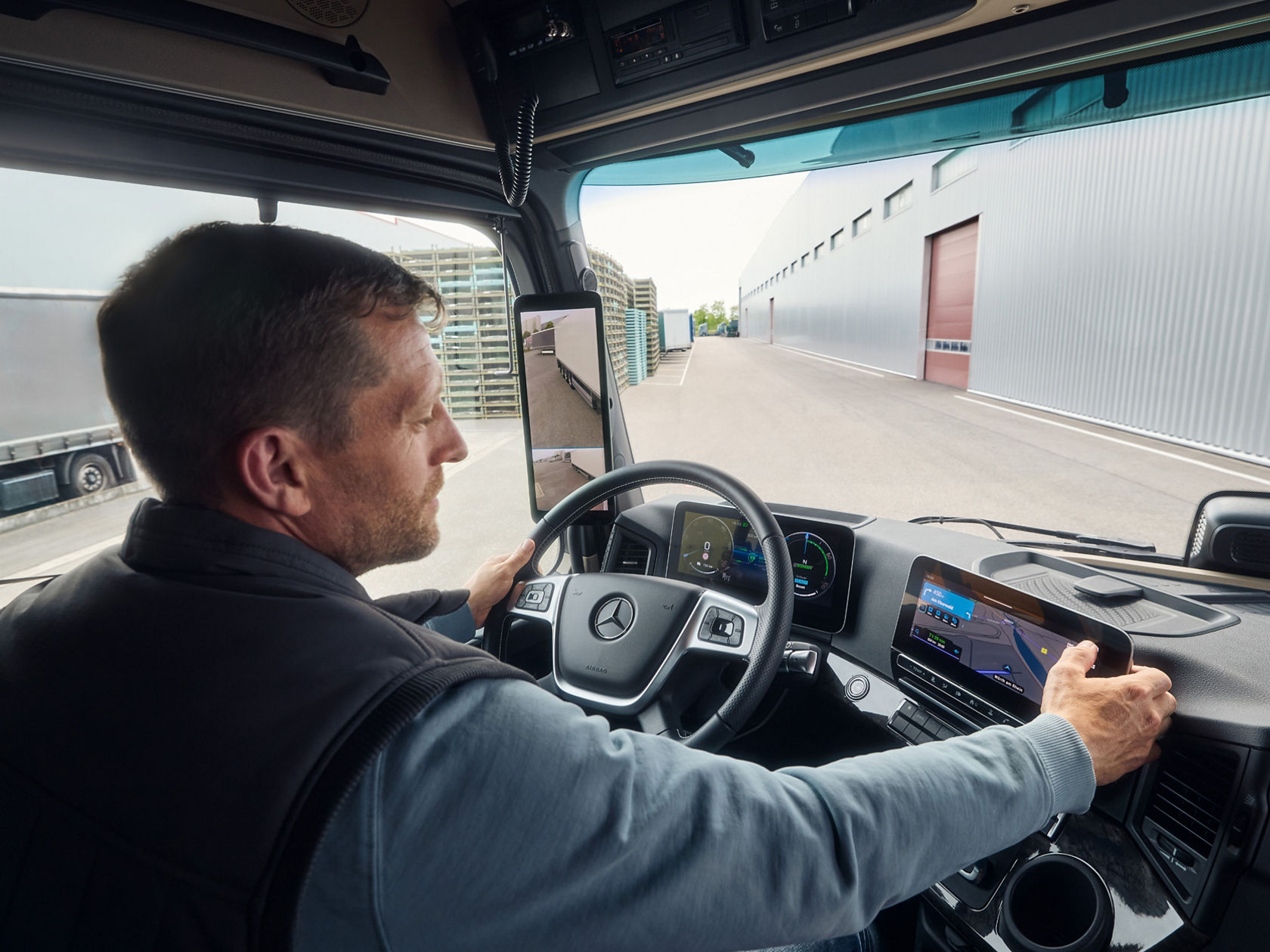 A man in work clothes sits at the wheel of an eActros from Mercedes-Benz Trucks, looking to the right while holding the steering wheel. The dashboard features digital displays and a large screen showing a map. Through the windscreen, you can see an industrial area with a large building.
