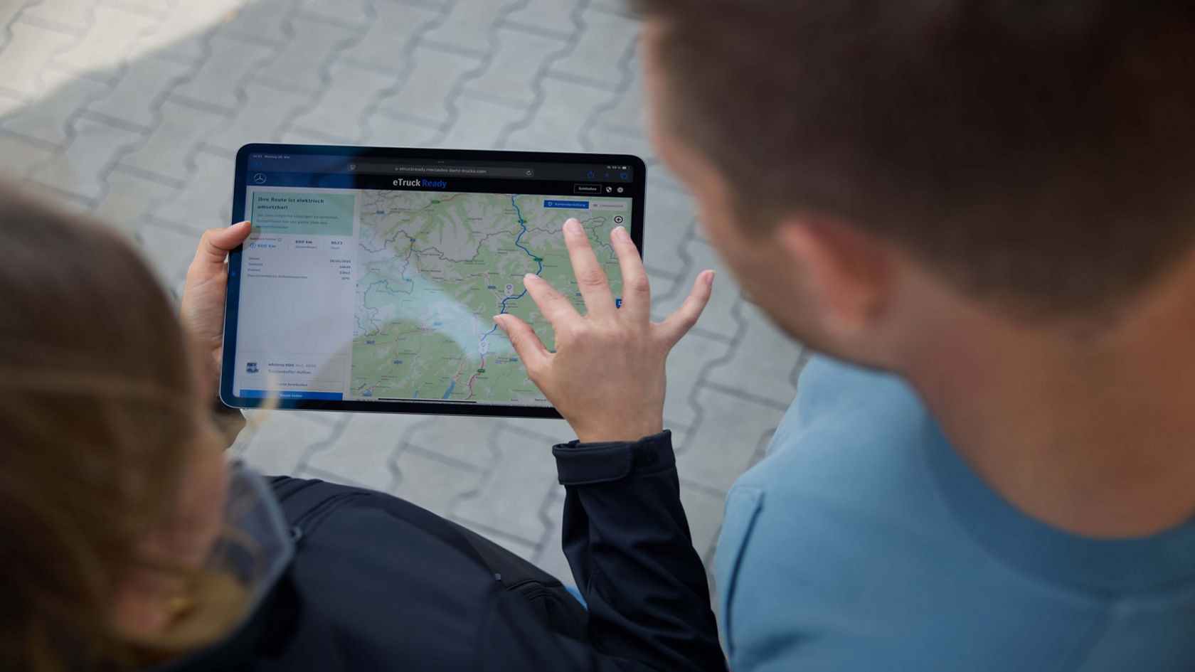 Two people, one of whom is holding a tablet and tapping it, look at a digital map together, which shows a route and topographical details. The scene is shot from above, putting the interaction with the tablet and the people’s heads in the foreground.