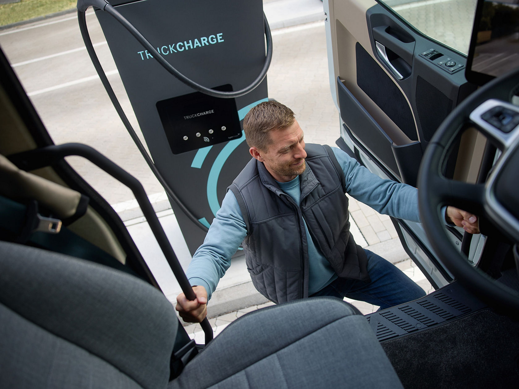 A man in work clothes climbs into the eActros from Mercedes-Benz Trucks; a TruckCharge charging station is visible in the background. The focus is on the driver’s entrance as he holds on to the grab handle.