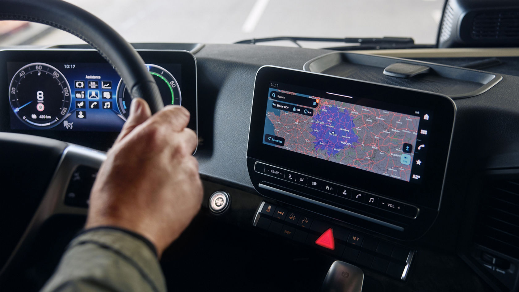 The image shows the modern cockpit of a truck from the driver’s perspective, with one hand holding the steering wheel. The focus is on the two large digital displays: the instrument cluster behind the steering wheel and the central infotainment screen, which shows a navigation map.