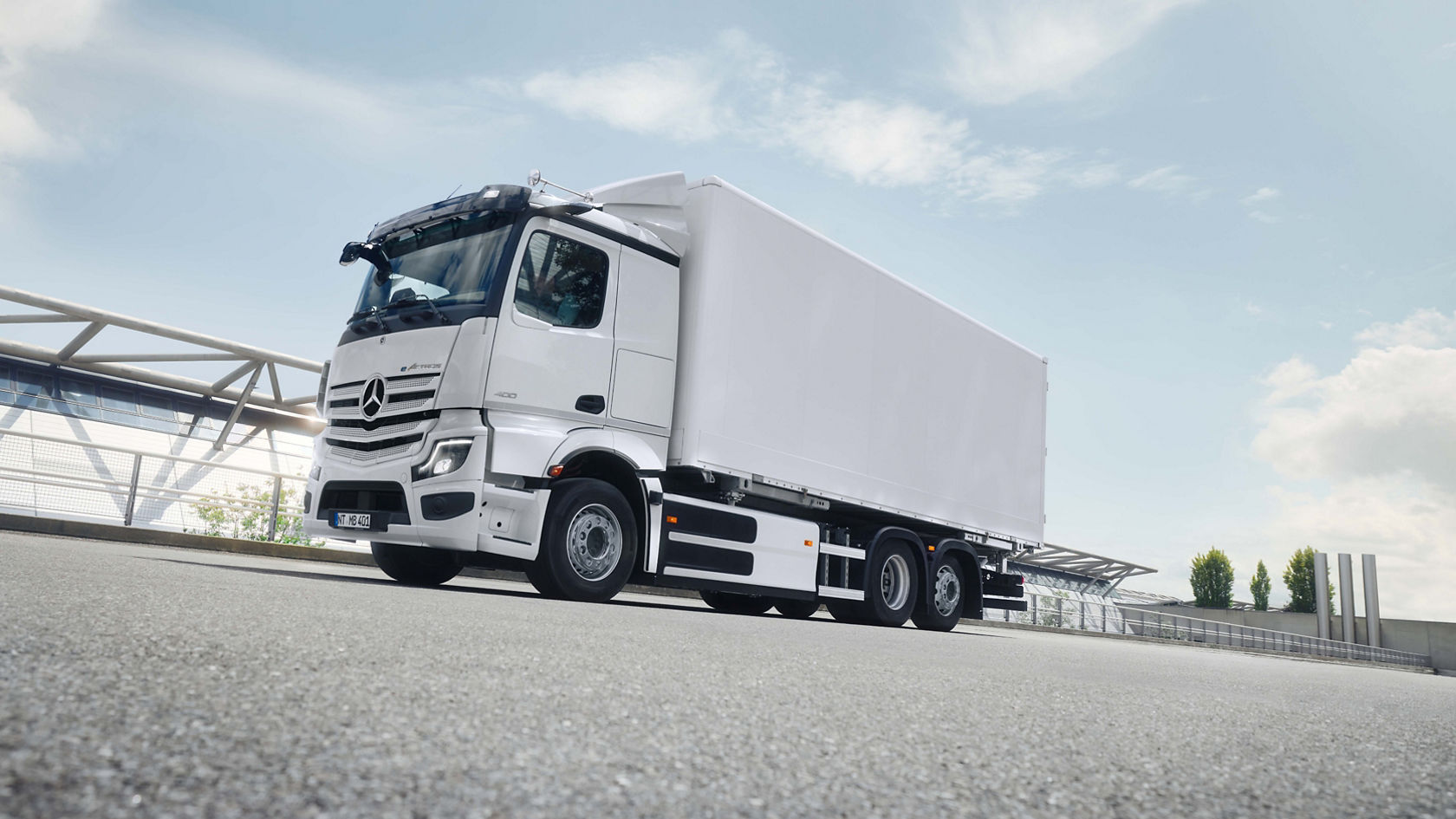 A white eActros 400 with a box body drives on a tarmac road under a partially cloudy sky. The lorry can be seen from the front in the bottom left of the picture and is moving towards the top right. In the background is a modern bridge construction made of metal struts and glass. The road is grey and has a slightly rough texture. The lighting suggests a sunny day.