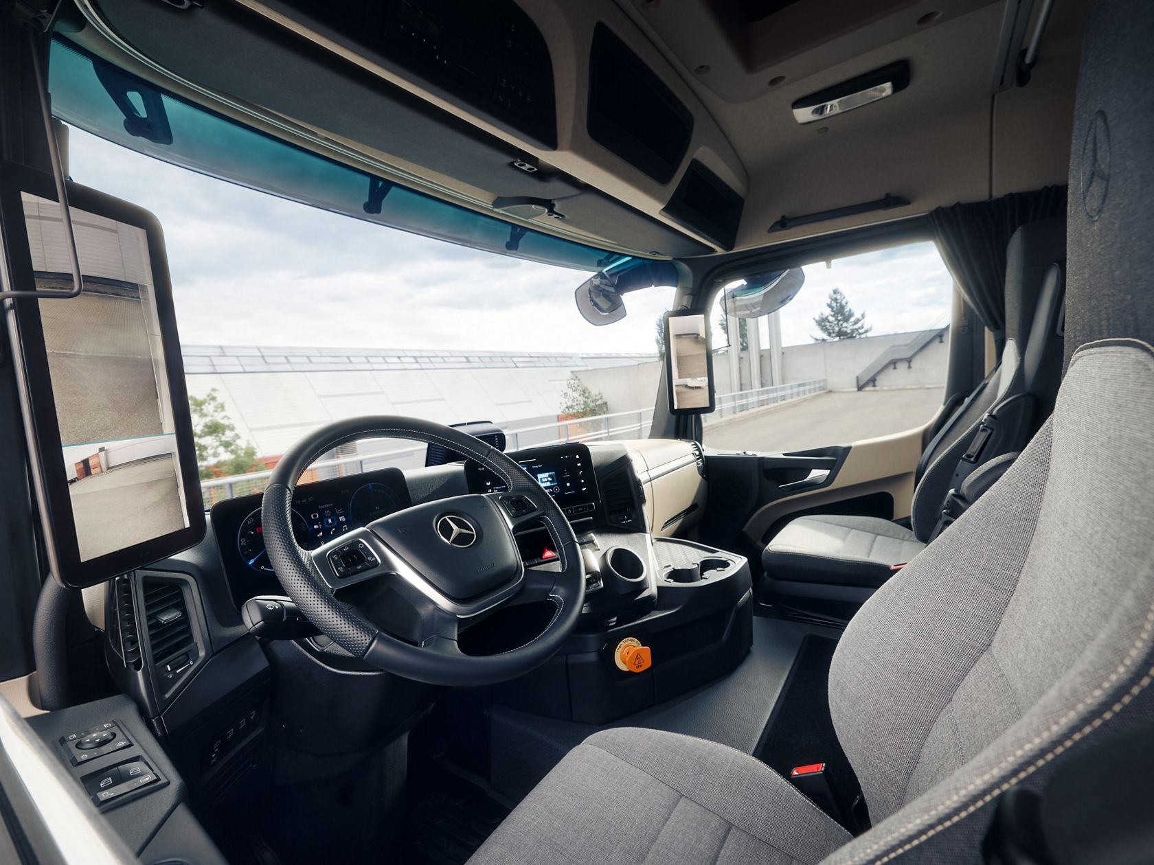The image shows the interior of the eActros from Mercedes-Benz Trucks from the perspective of the front passenger seat. You can see the steering wheel with the Mercedes star, the dashboard with digital displays and screens, as well as the comfortable seats in light grey fabric.