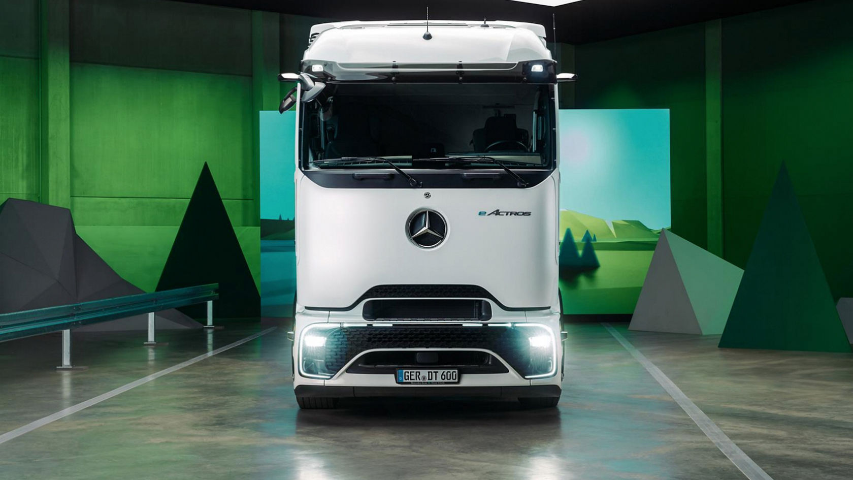 A white eActros from Mercedes-Benz Trucks stands facing forwards in a room with green walls and geometric shapes that represent a stylised landscape. The floor reflects the light and the surroundings, presenting a modern and stylistic view of the truck.