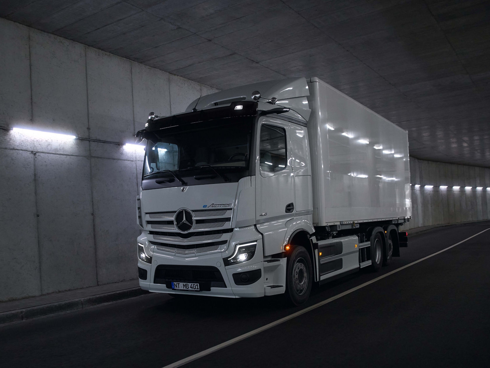 A white eActros from Mercedes-Benz Trucks with a box body drives through an illuminated tunnel. The lights on the tunnel walls reflect off the shiny surface of the truck, creating a dynamic scene.
