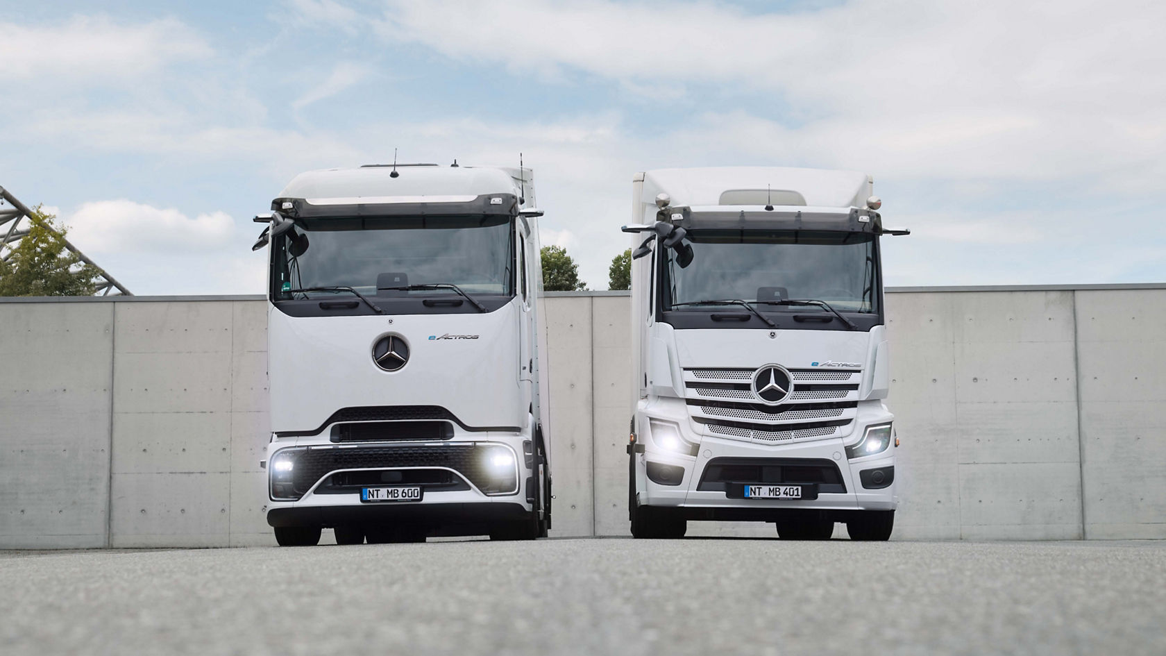 Two white eActros from Mercedes-Benz Trucks, one with ProCabin and one with the tried-and-tested cab, stand on tarmac in front of a large building with roller doors. The left truck has a semitrailer, while the right truck has a box body. Both trucks are clean and modern, and the sunlight is reflected in their windscreens.