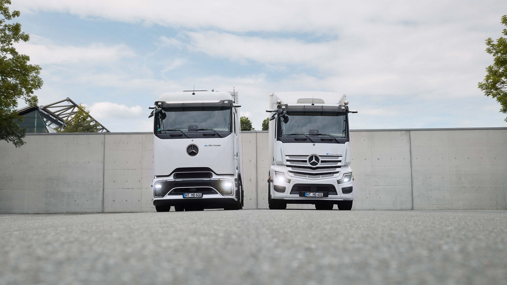 Two white eActros from Mercedes-Benz Trucks, one with ProCabin and one with the tried-and-tested cab, stand next to each other on tarmac in front of a concrete wall. Both trucks have their headlights on; trees and a modern building can be seen in the background.