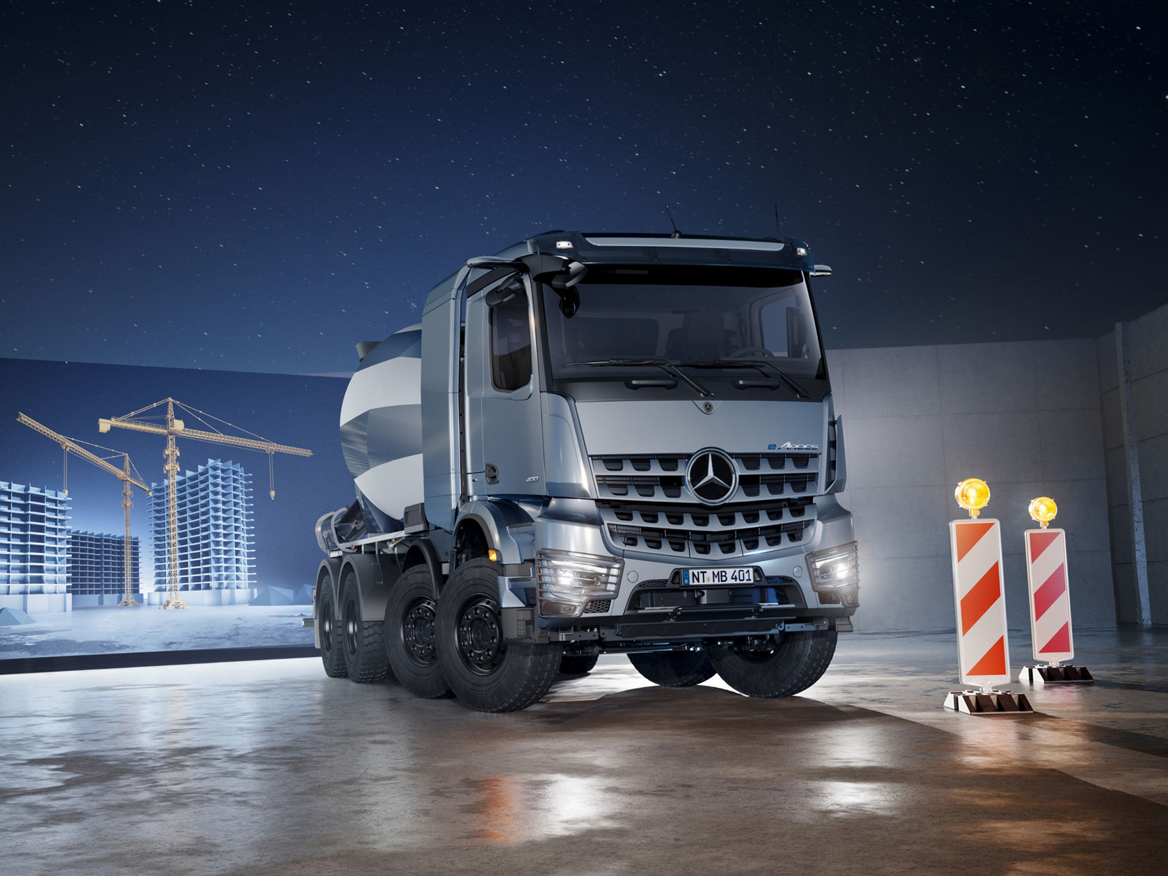 The image shows a blue-grey Mercedes-Benz eArocs concrete mixer standing in a hall. In the background, an LED wall shows a simulated scene of a construction project at night with illuminated cranes and bodyshells under a starry sky. In front of the truck are two red and white-striped warning beacons with yellow flashing lights.