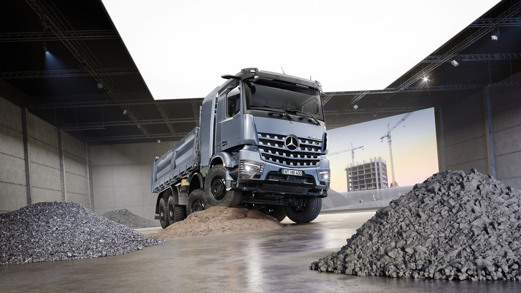 The image shows a blue-grey eArocs 400 tipper standing on a small hill of sand in a large hall. There are large piles of gravel to the left and right of the truck. In the background is an LED canvas with the image of a construction site.