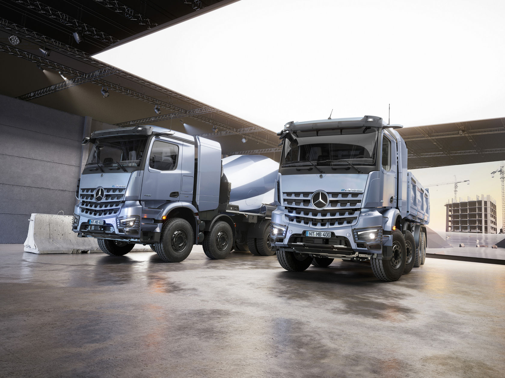 The image shows two eArocs 400 in a large hall with concrete floor and lighting on the ceiling. The eArocs 400 on the right is a concrete mixer, while the eArocs 400 on the left is a tipper. In the background, a large LED screen displays an image of a construction site.