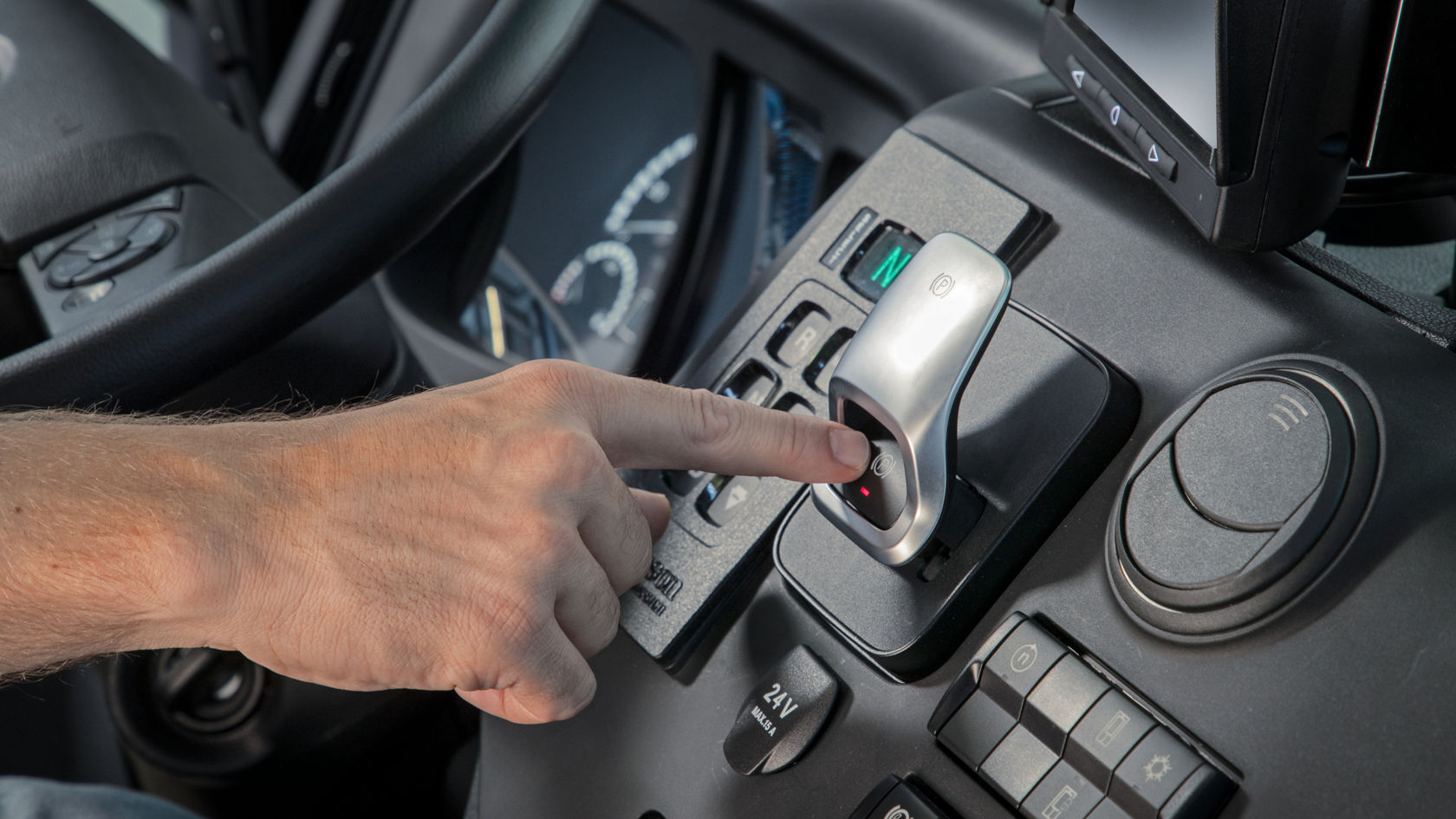A hand presses a button on an electronic gearshift lever. The gearshift lever is located on the centre console and is surrounded by other controls and displays.