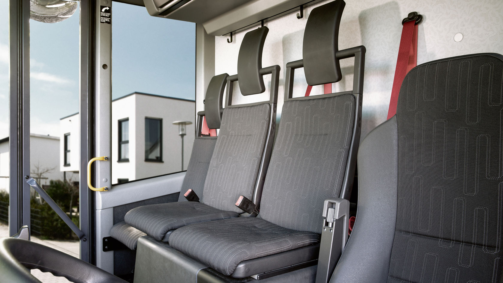Close-up of three empty passenger seats. The seats are in grey fabric with black head restraints and red seat belts.