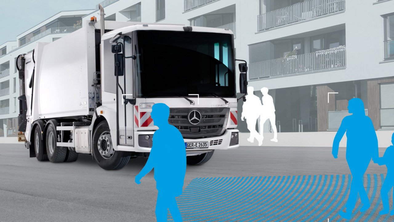 A silver Econic in municipal use for waste disposal stands in front of a modern residential building. Blue silhouettes of pedestrians can be seen. Blue lines show the functional range of a safety assistance system.