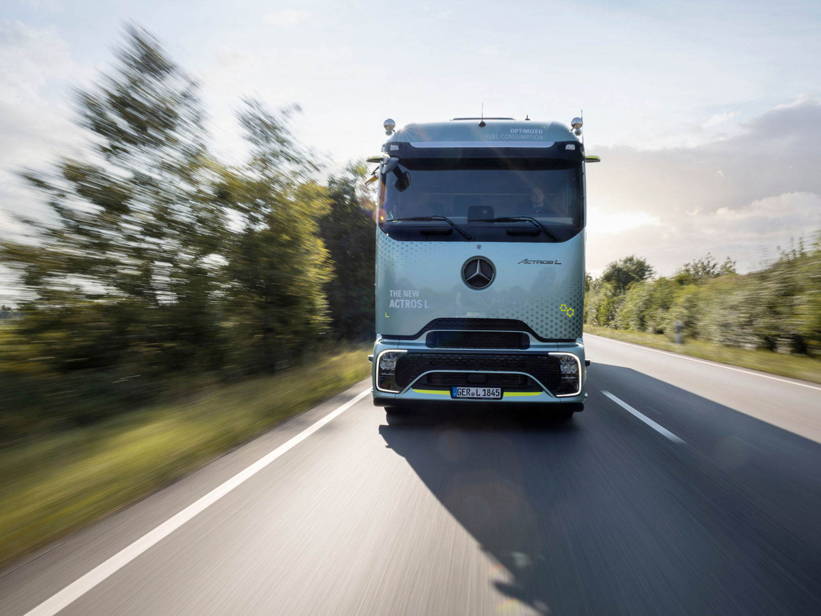 A mint-green Mercedes-Benz Actros L with ProCabin drives on a road lined with trees. It is shown from the front. The sun shines brightly and casts a long shadow of the lorry on the asphalt.