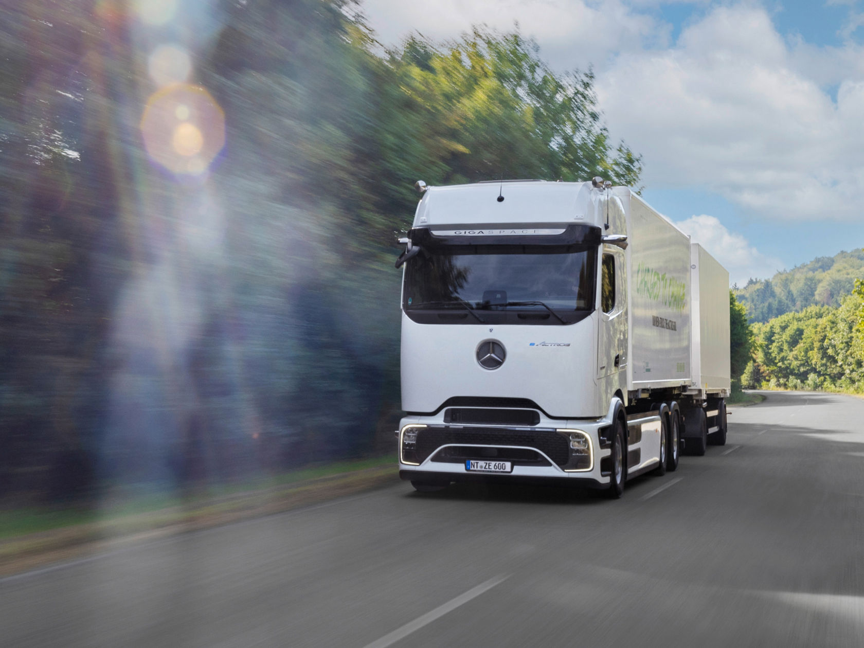 A white truck drives on an asphalted road through a wooded landscape. The truck can be seen from the front left and is moving towards the right. The trees on both sides of the road are blurred, indicating movement. There are some clouds in the sky. The sun shines from the top left and creates a glow in the image.