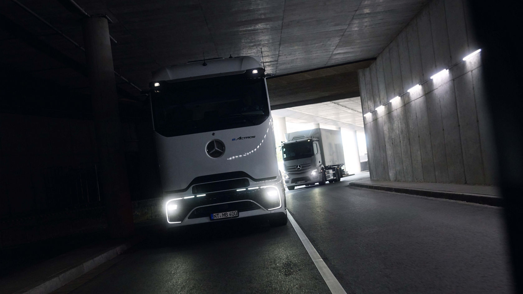 Two eActros 400, one with the ProCabin and one with the tried-and-tested cab, drive through a dark tunnel with illuminated headlamps. Bright daylight can be seen at the end of the tunnel, contrasting with the dark surroundings.