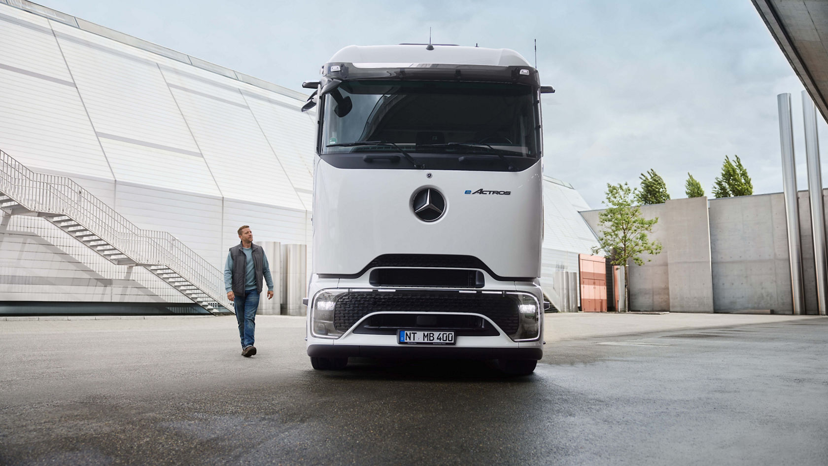 A man in work clothes walks on tarmac; a white eActros 400 with ProCabin from Mercedes-Benz Trucks is parked in the background. On the left is a modern building with a long outside staircase and on the right, trees and other building structures.
