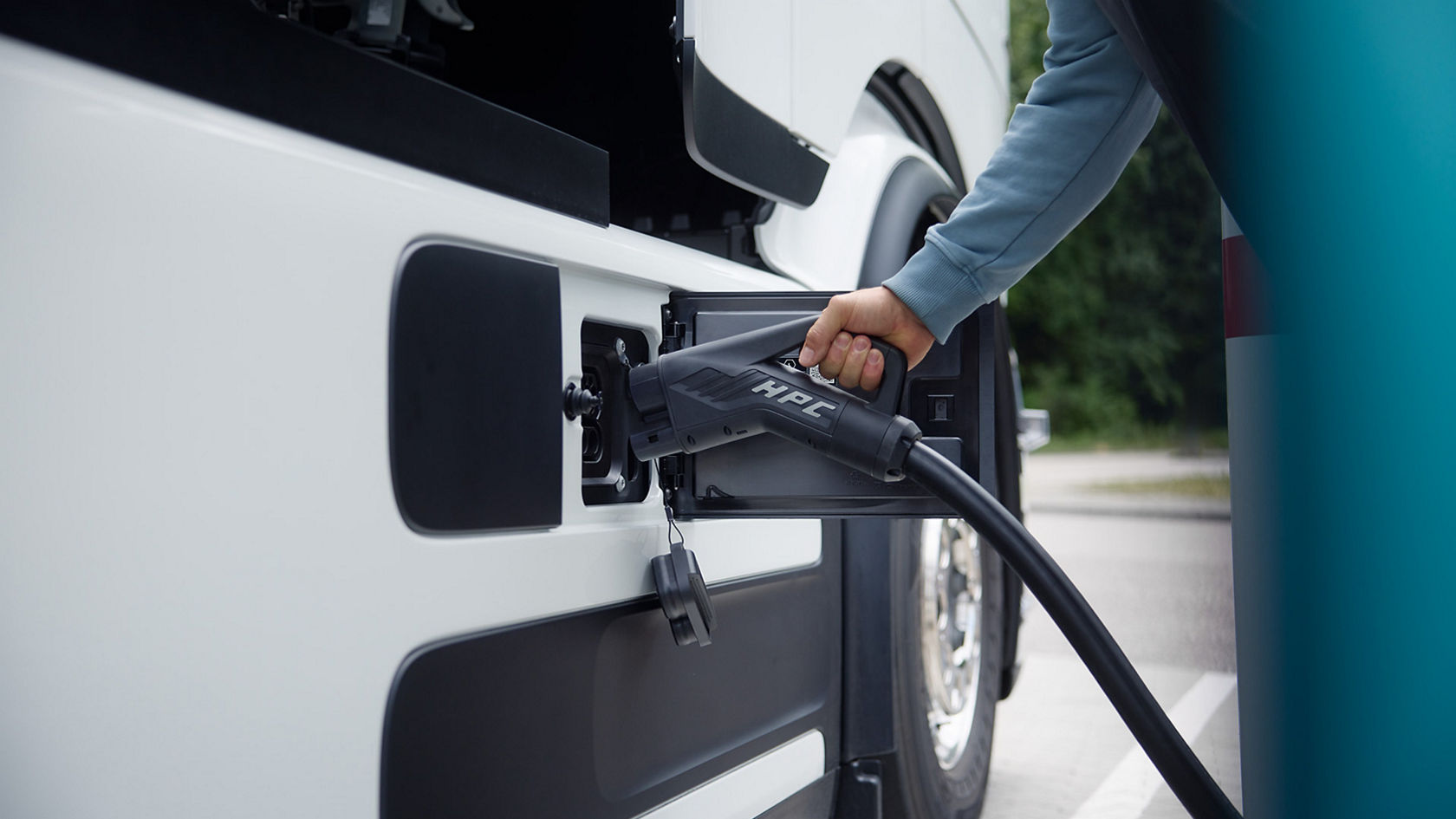 A person plugs a charging cable into the eActros from Mercedes-Benz Trucks, which is white and partly visible in the picture. The focus is on the charging process, with the cable and connector clearly visible.