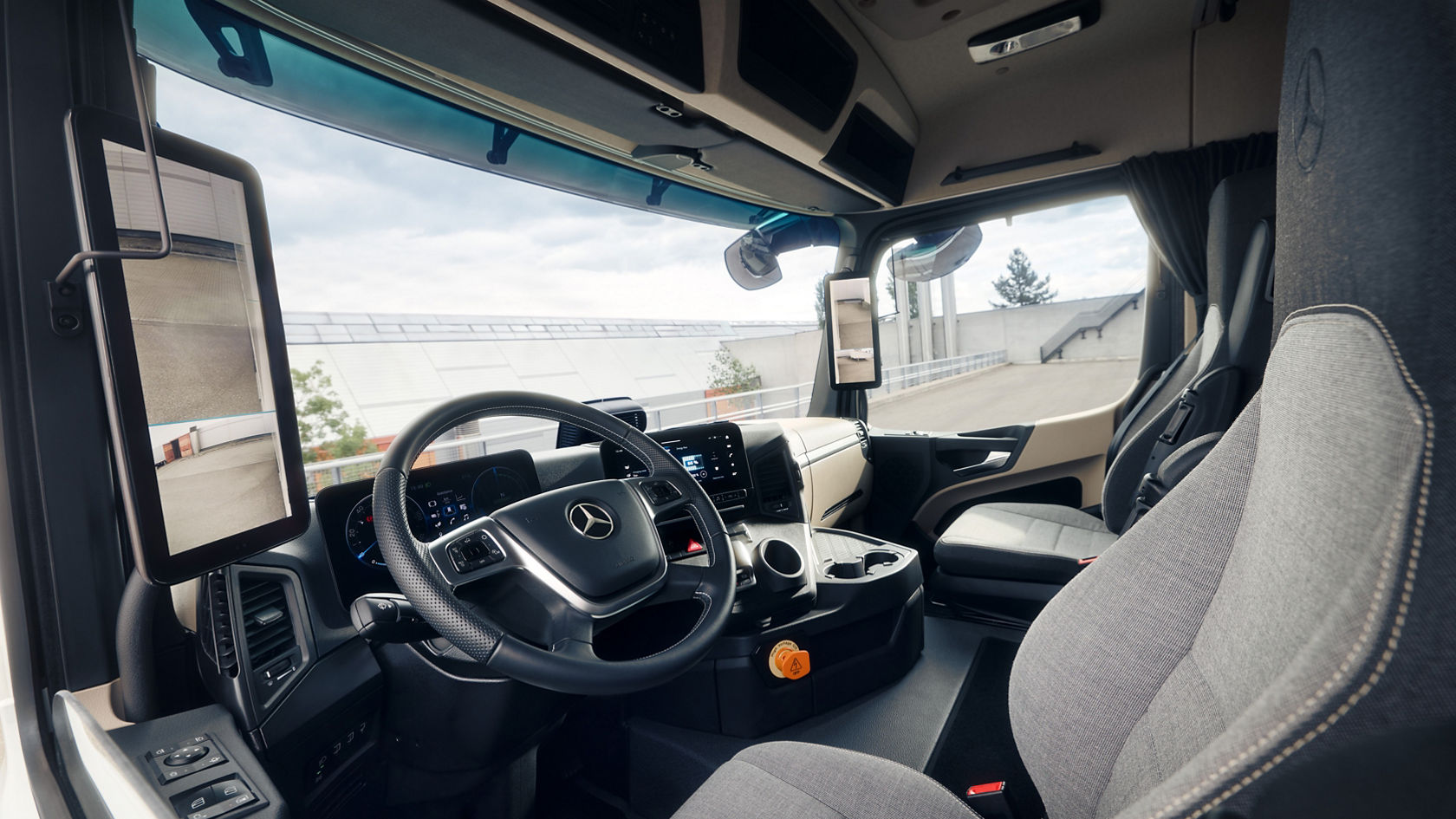 The image shows the interior of the eActros from Mercedes-Benz Trucks from the perspective of the front passenger seat. You can see the steering wheel with the Mercedes star, the dashboard with digital displays and screens, as well as the comfortable seats in light grey fabric.