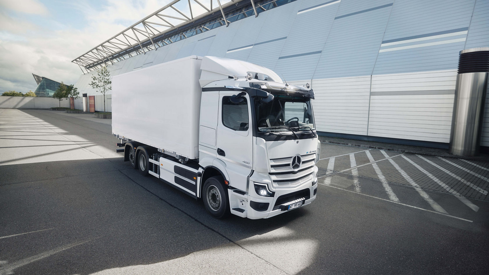 A white eActros 400 from Mercedes-Benz Trucks with a box body stands in a tarmac car park in front of a modern building with a glass façade. The sunlight casts long shadows on the ground, indicating a clear day.
