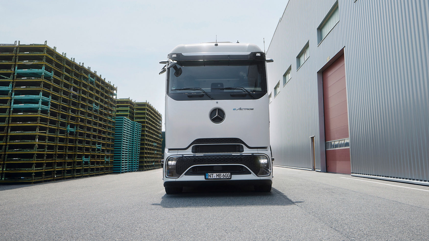 An eActros 600 with ProCabin from Mercedes-Benz Trucks stands facing forwards on tarmac. On the left side of the picture are high stacks of yellow and blue pallets, while on the right side is a large, modern building with a grey corrugated sheet metal façade and a red door. The sky is bright and slightly cloudy.