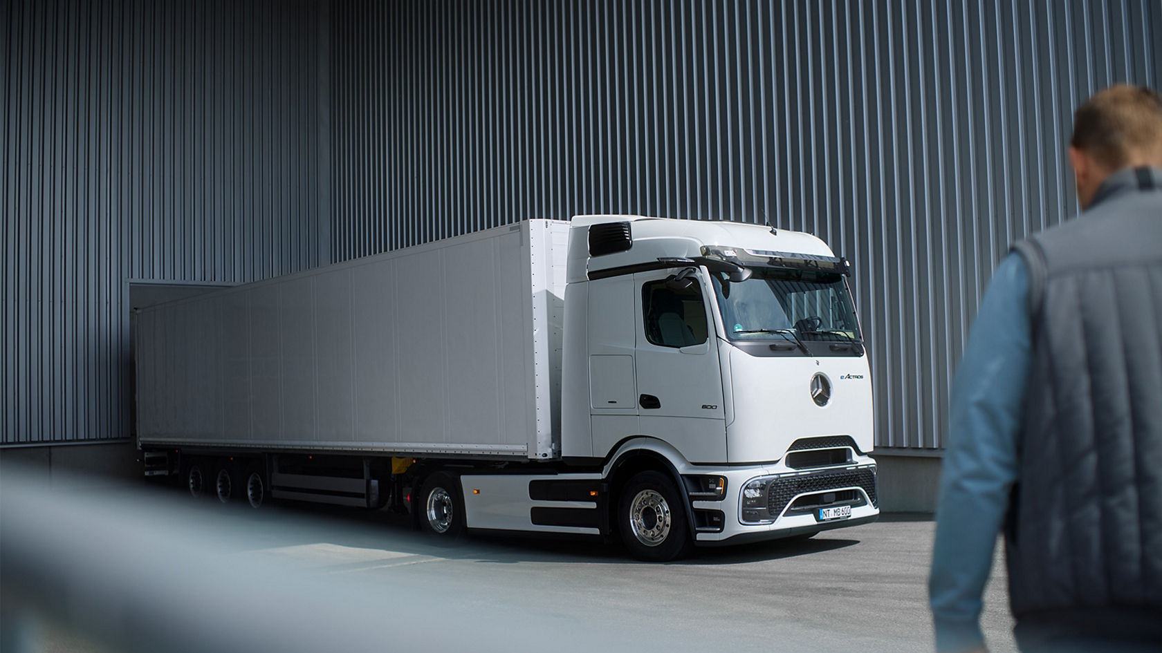 A white eActros 600 with ProCabin from Mercedes-Benz Trucks with a long, white trailer stands on tarmac in front of a grey corrugated sheet metal wall. The truck can be seen from the side, with the front pointing slightly to the left. In the foreground, with their back to the observer, a person in a blue jacket and a grey vest is looking at the lorry. The scene is lit from above.