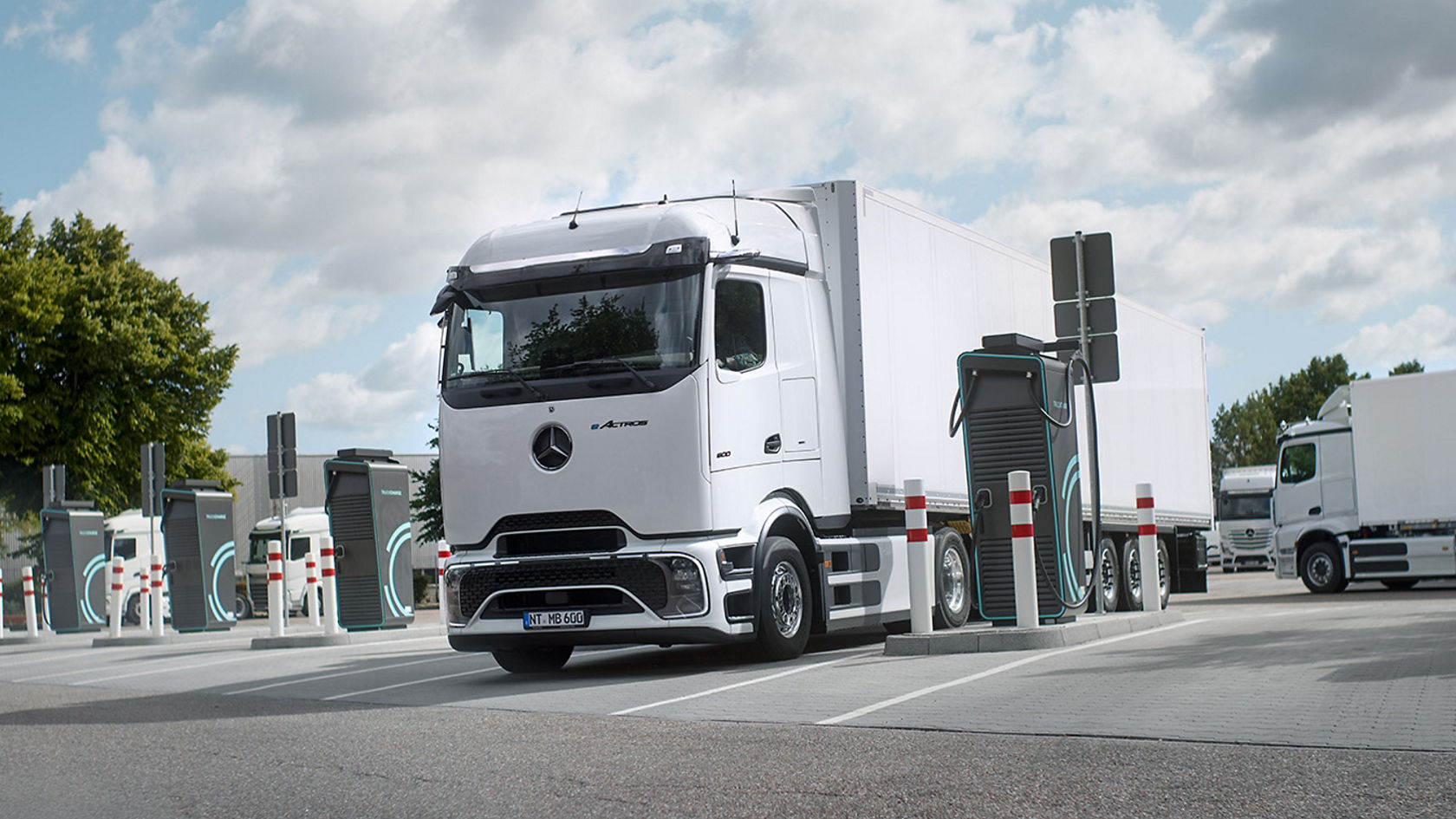 A white eActros 600 with ProCabin from Mercedes-Benz Trucks with a white trailer is parked in a car park and is being charged at a charging station. Several other charging stations and barrier posts can be seen around the truck. There are other white lorries in the background, and green trees can be seen on the left side of the image under a partially cloudy sky.