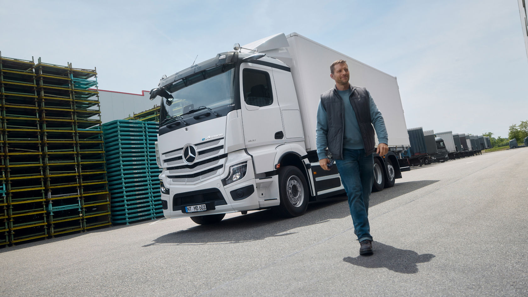 A man in work clothes walks on tarmac; a white eActros 600 from Mercedes-Benz Trucks with a trailer is parked in the background. On the left are stacked pallets and on the right, a large building that may be a warehouse or industrial complex.