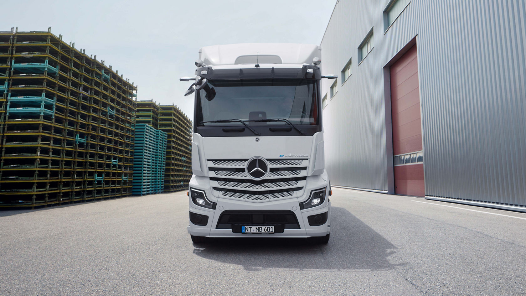 An eActros 600 from Mercedes-Benz Trucks stands on tarmac in front of a modern building with a long outside staircase. The truck is white and has the Mercedes star logo on the radiator grille, while trees and other building structures can be seen in the background.