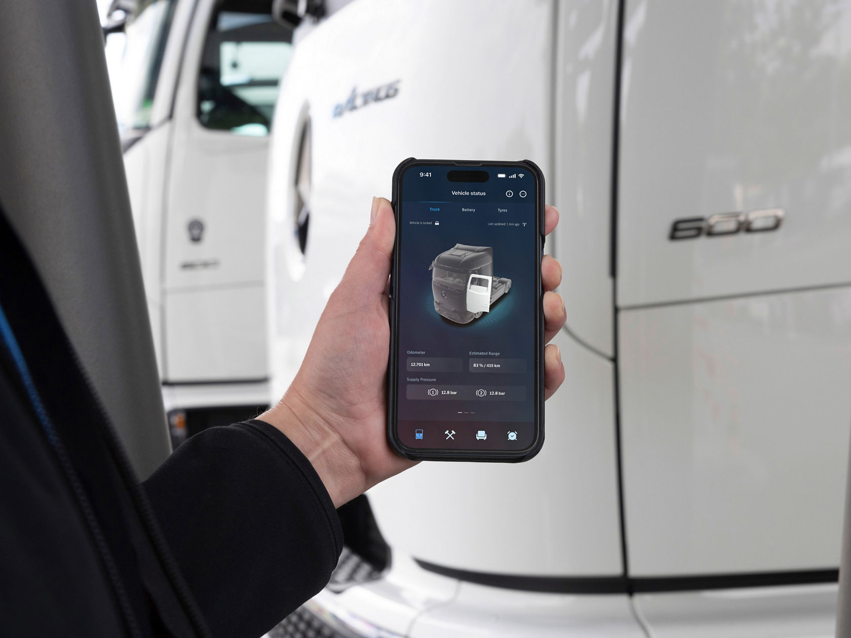 A hand holding a smartphone showing an app that displays the status of an eActros from Mercedes-Benz Trucks, including kilometre reading and estimated range. In the background is the white truck, illustrating the connection between the app and the vehicle.