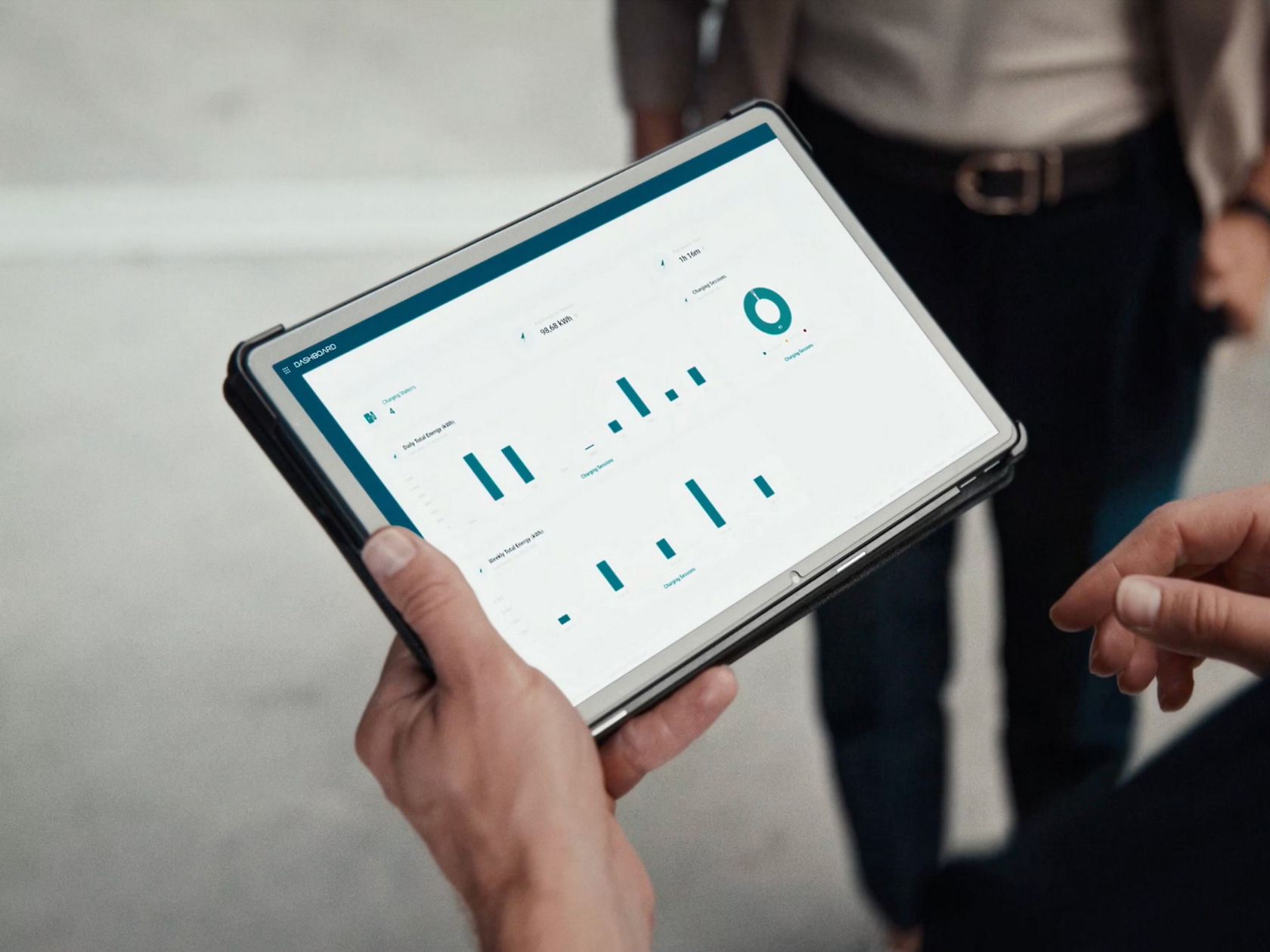 A person holds a tablet with a dashboard displaying various graphs and data for energy management and charging operations. The legs of another person can be dimly seen in the background.