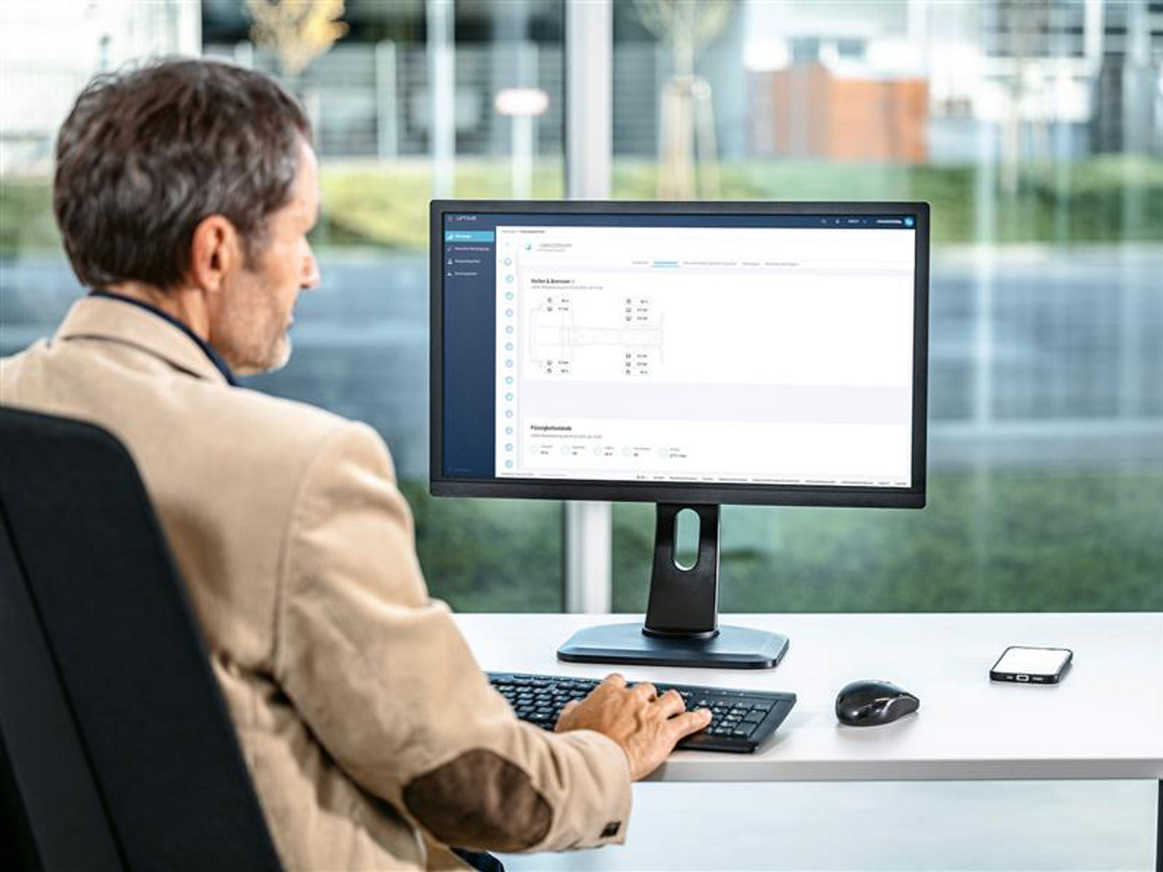 A man sits in front of a computer monitor and types on a keyboard with the screen showing a software interface with graphs and data. In the background, trees and buildings are blurred through a window, indicating an office environment.