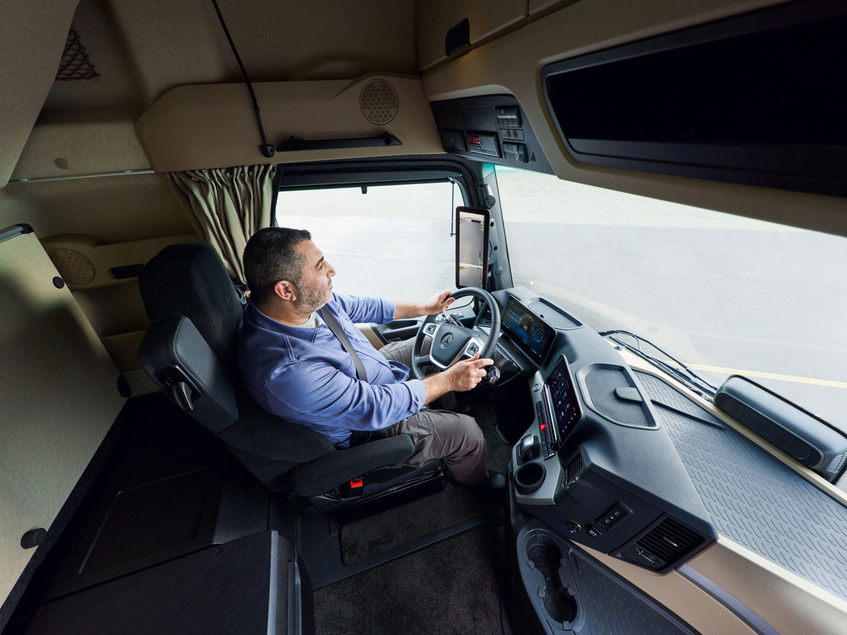 A man with a beard and a blue shirt sits in the driver’s seat of an Actros L, holding the steering wheel. He looks to the right, while the road in front of him is visible through the windscreen.