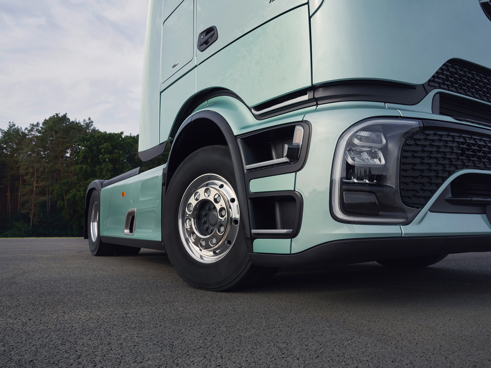 A large, mint-green Actros L standing on an asphalt surface, with the focus on the front end and the front wheel. Trees and a cloudy sky can be seen in the background.