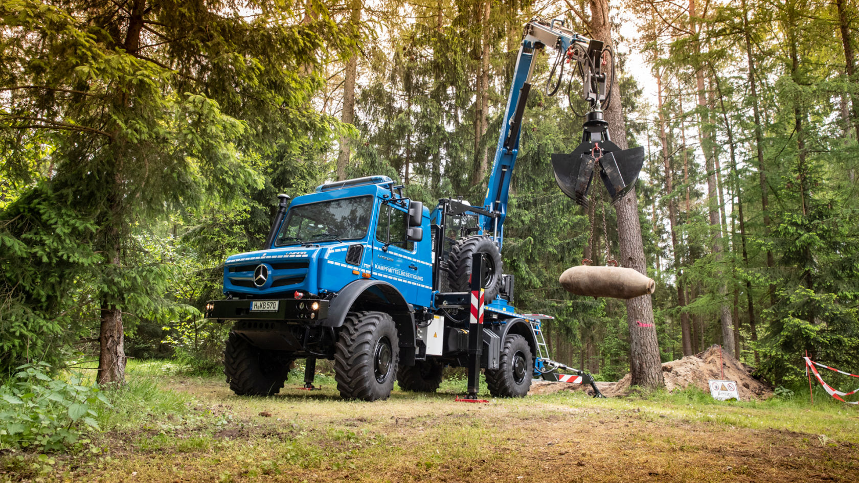 A blue Unimog with a crane arm lifts a large cylindrical load in a forest area.