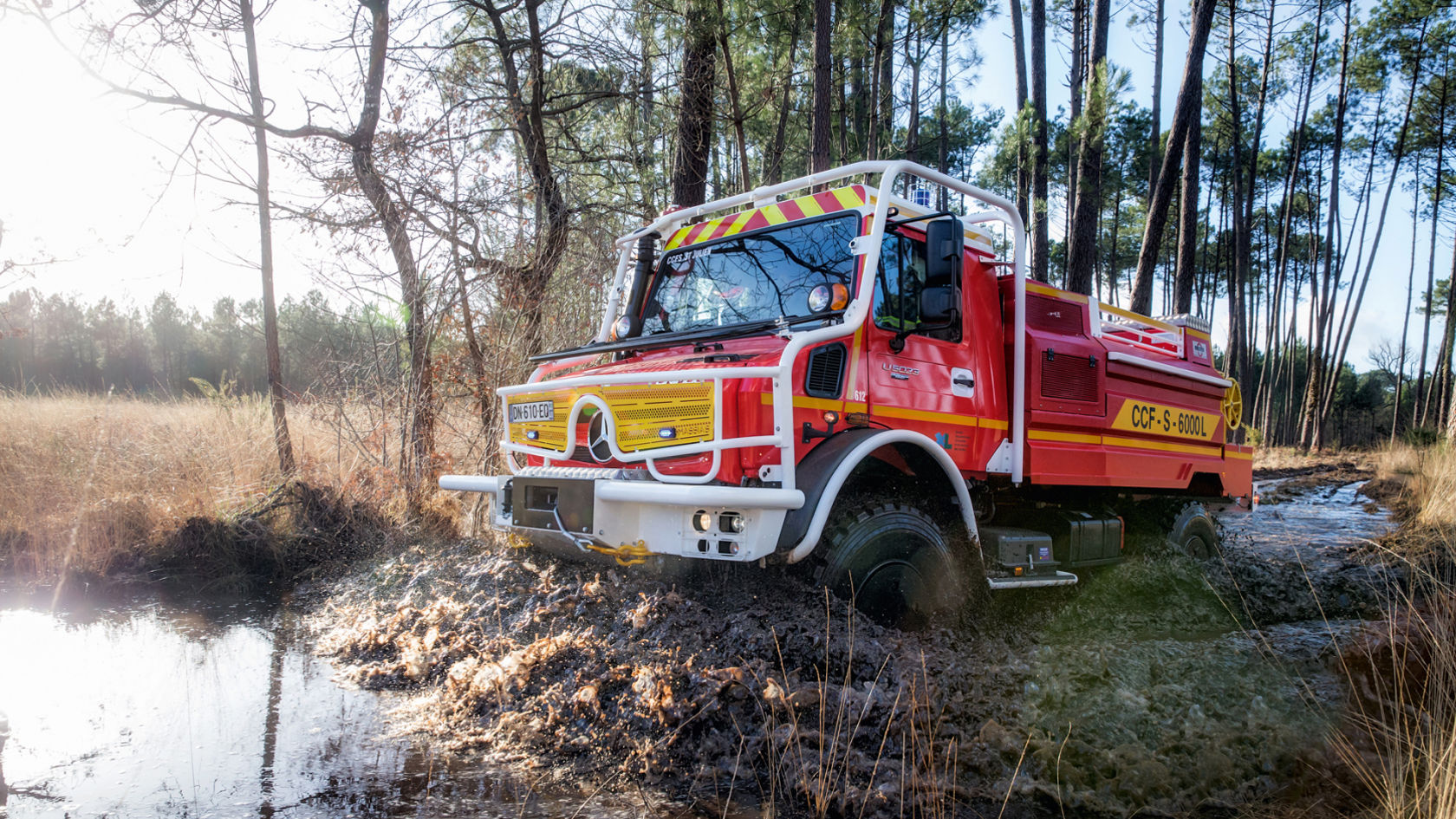 A red-yellow Unimog fire truck drives through muddy water in a forest area.