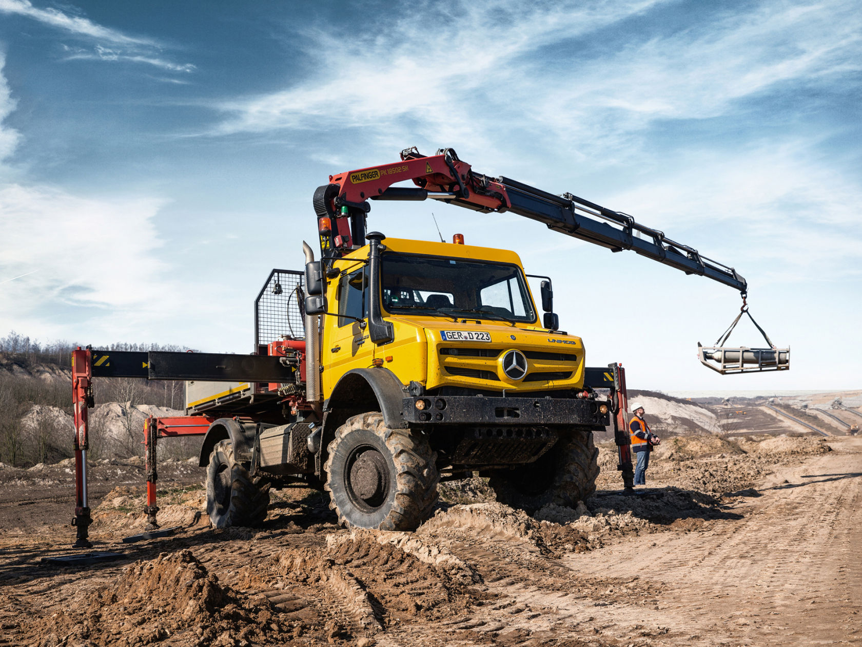 A yellow Unimog with a crane lifts a load in a quarry or similar environment. A person in work clothing standing next to the vehicle.