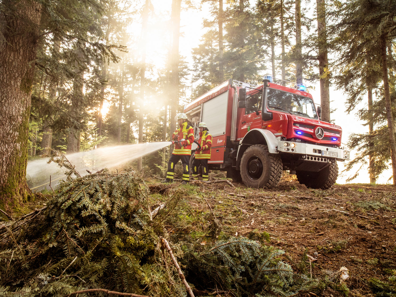 Red fire truck standing in a forest, while emergency workers spray water with a hose.