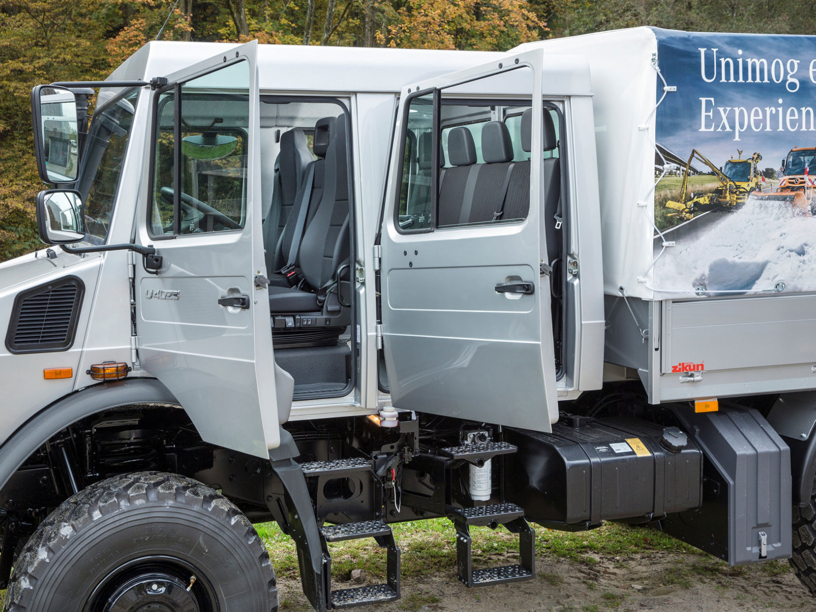 A silver Unimog with open doors, showing the interior and seats. The cargo area of the lorry is covered with a tarpaulin.