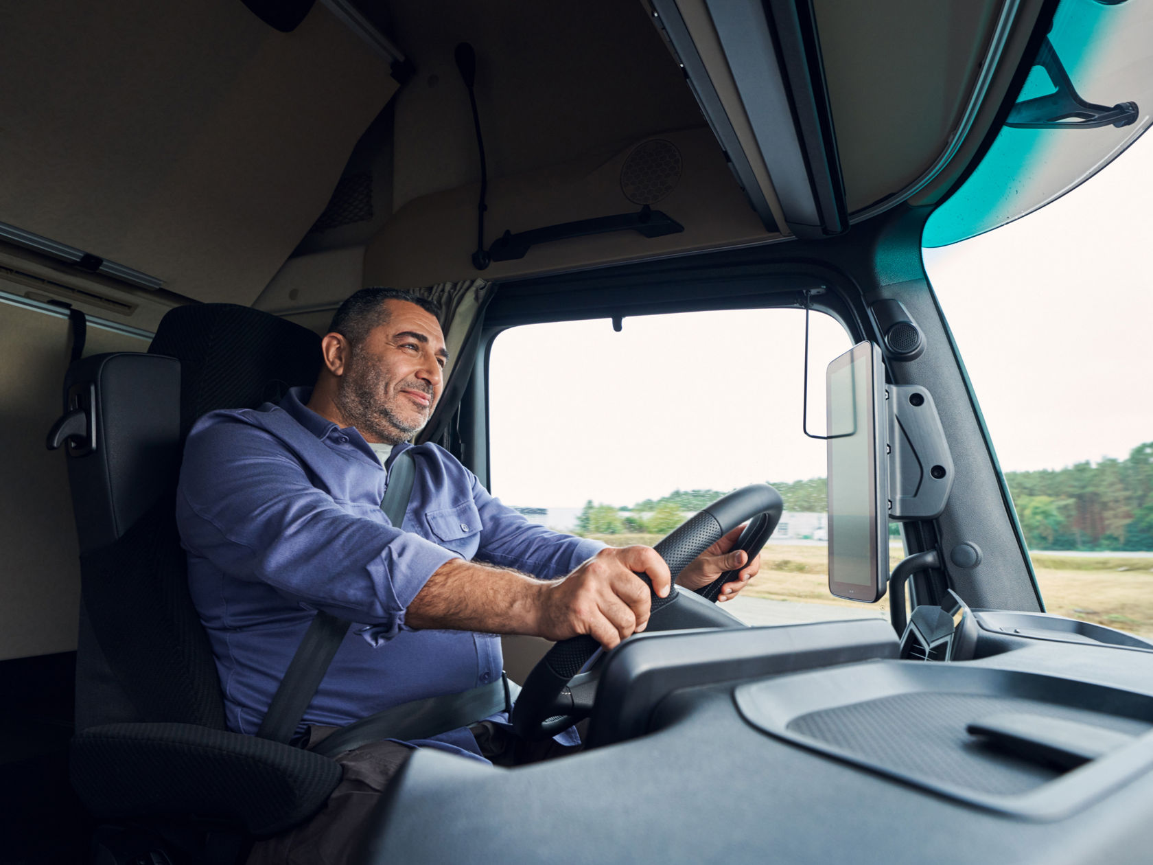 A man with a beard and a blue shirt sits behind the wheel of an Actros L, smiling. He is holding the steering wheel with both hands while driving on a road bordered by trees.