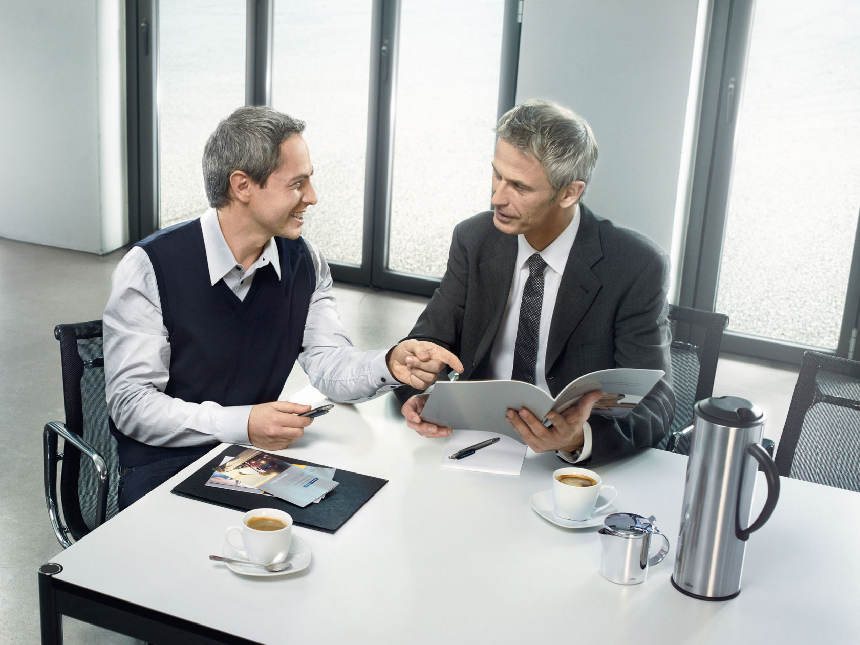Two businessmen sitting at a table, discussing documents and drinking coffee.