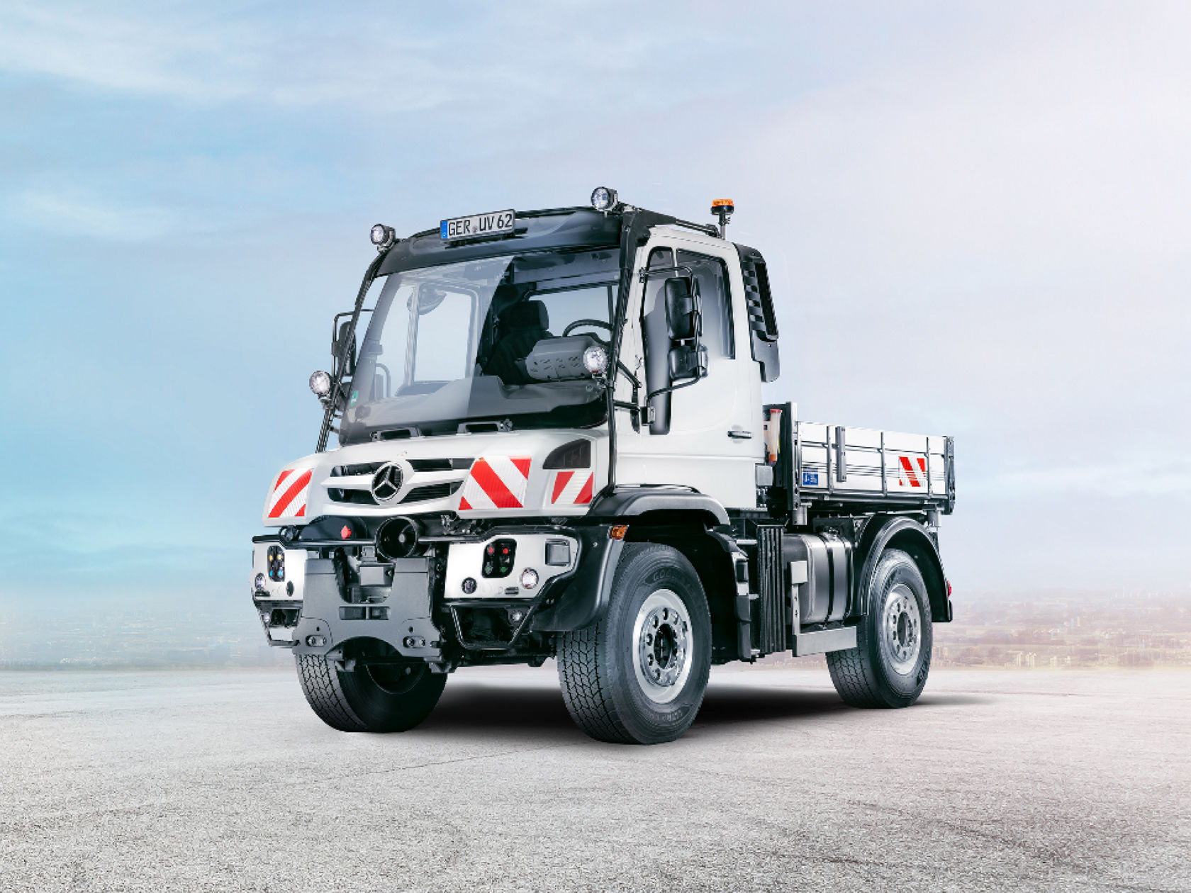 A white Unimog stands on an empty road. The truck has an open cargo area and a large cab.