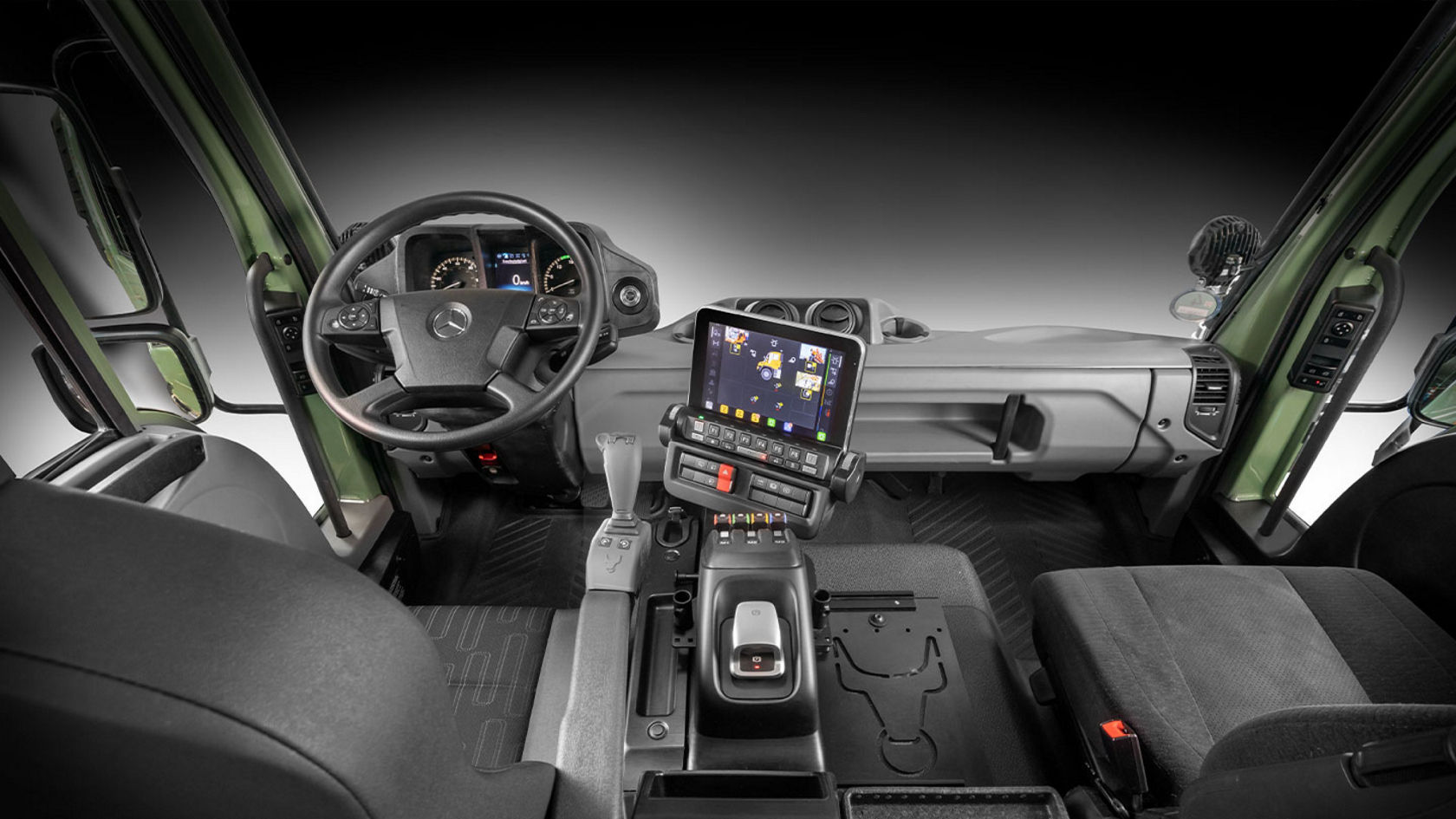 The interior of a vehicle cab with steering wheel, dashboard and screen. The seats are black and the dashboard is greenish-grey.