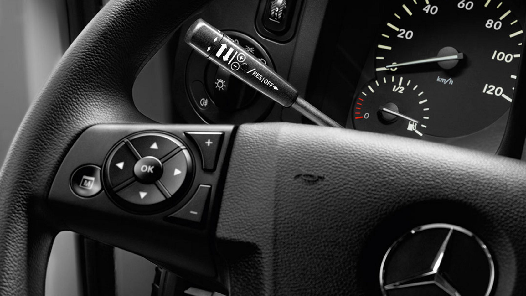 Close-up of a steering wheel with various buttons and controls. The dashboard with speedometer and rev counter can also be seen.