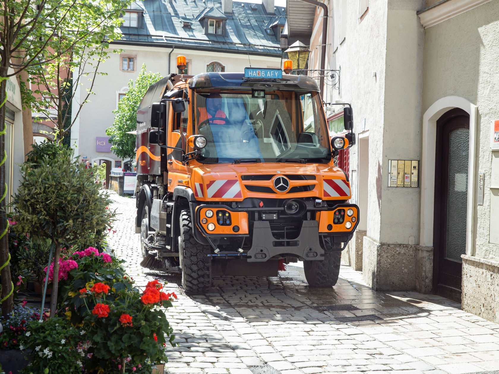 An orange Unimog implement carrier in municipal operations drives through a narrow cobblestone alley. Flowerbeds can be seen on the left.