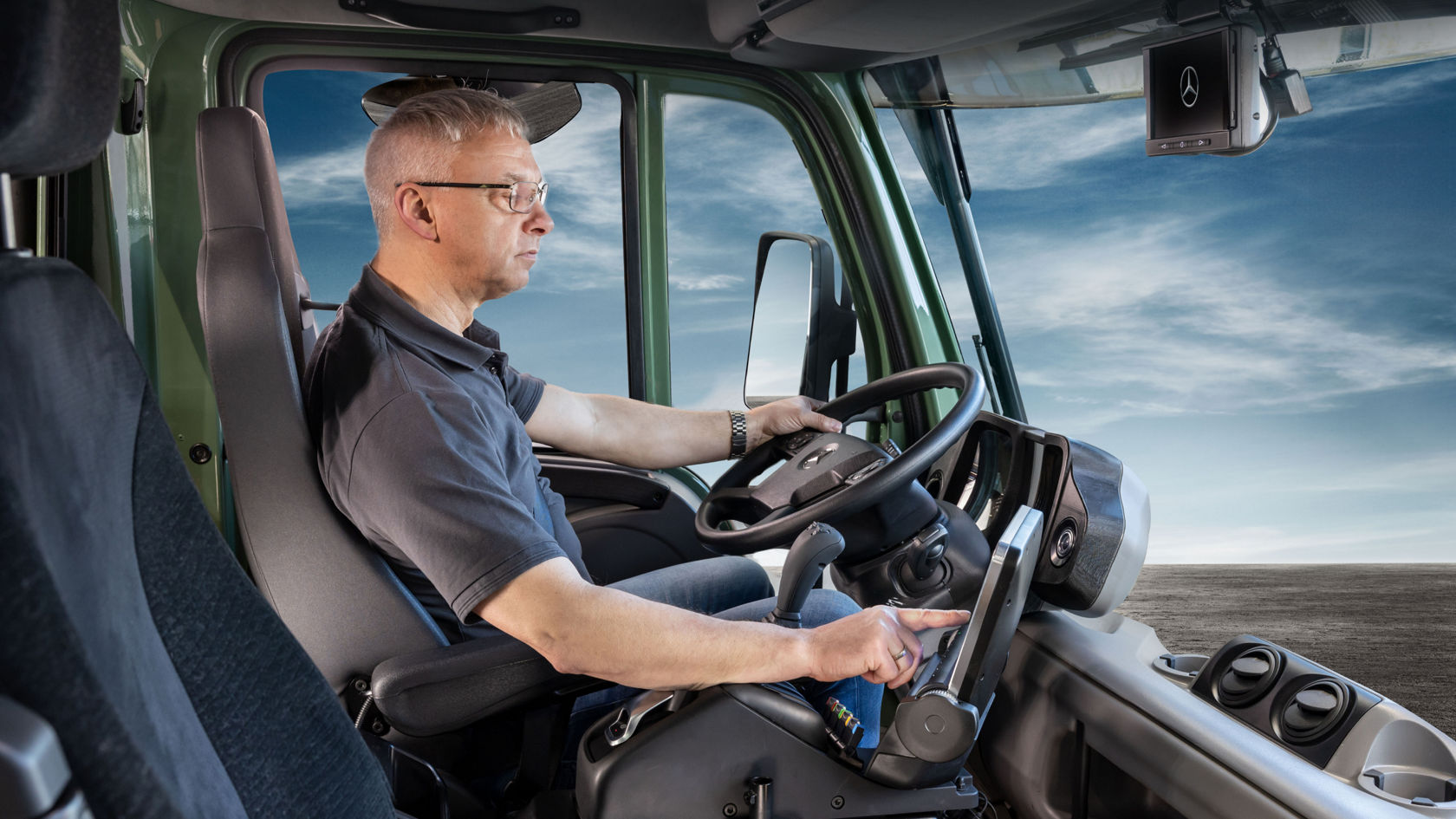 A man sits at the wheel of a Unimog and operating a control panel. He is wearing a blue shirt and glasses.