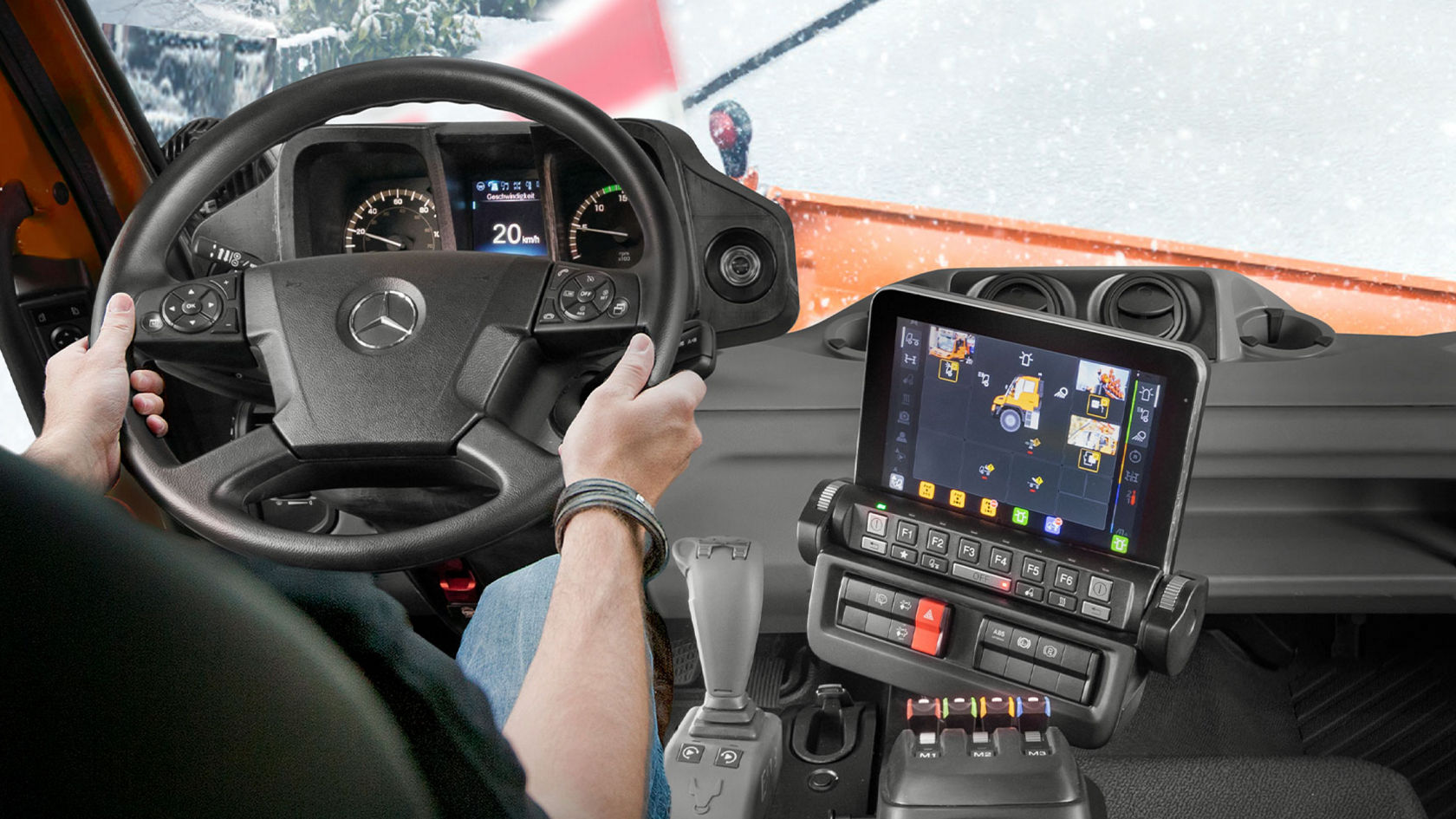 The view from the driver’s perspective of a Unimog implement carrier with a screen showing several camera perspectives. The driver has their hands on the steering wheel and a snow-covered road lies in front of them.