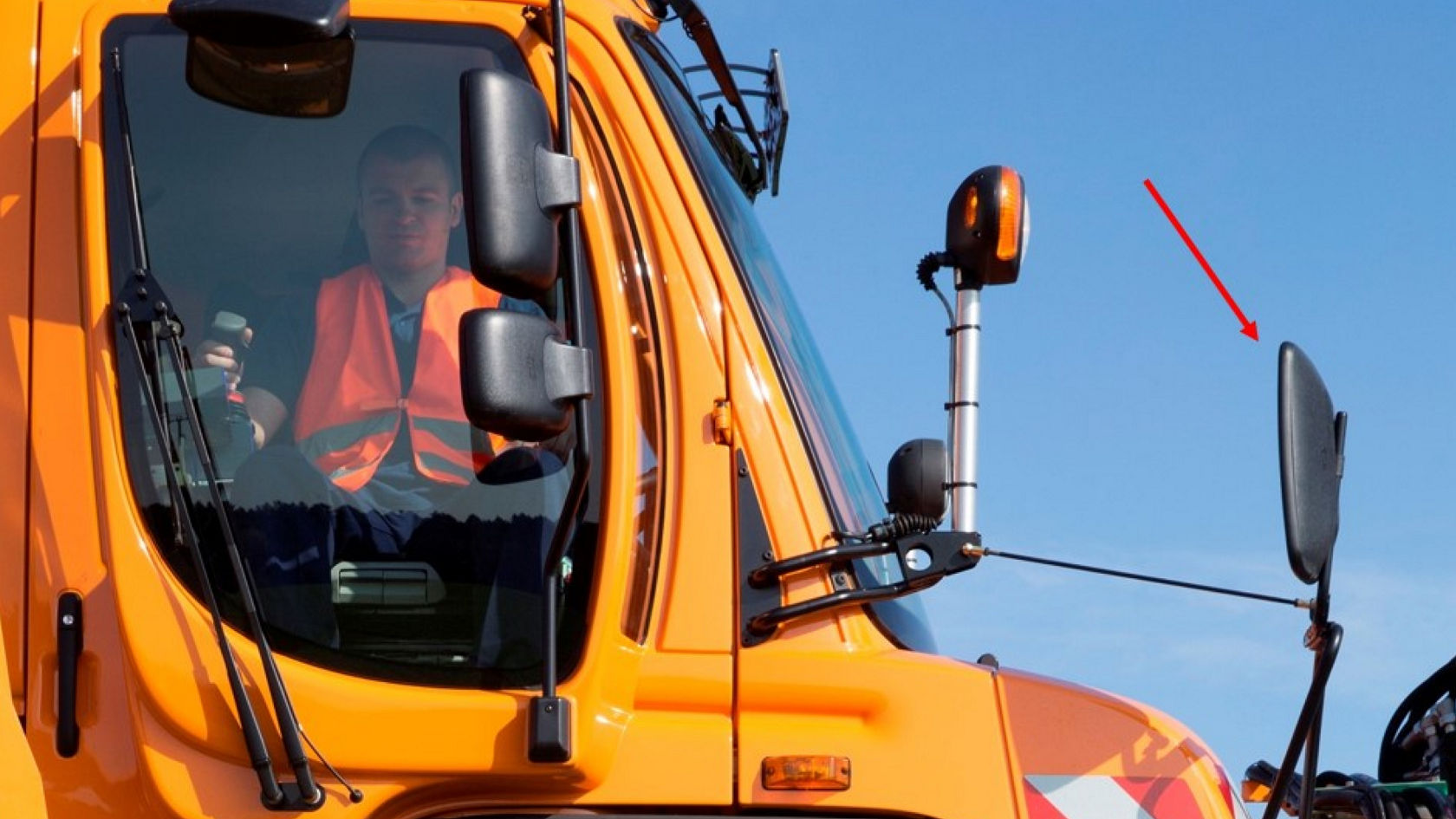 The side of an orange Unimog implement carrier. A driver wearing a high-visibility safety vest can be seen in the cab.